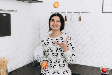 Cheerful woman standing in bright kitchen smiling laughing eating toss up ripe red apple apple...