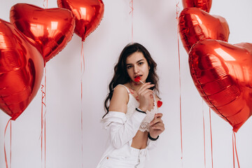 A smiling woman with kisses lipstick is holding eat candies and standing amidst heart-shaped helium balloons, celebrating a special holiday Valentines Day
