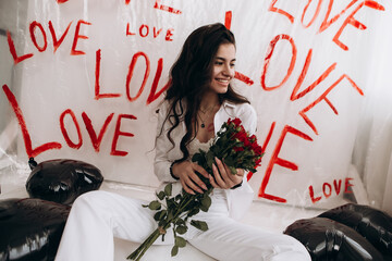 Woman sitting with kisses lipstick hold bouquet of red roses, black foil balloons, the word LOVE hand-painted on plain white surface oilcloth, Valentine`s day concept celebration decoration

