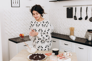 Cheerful woman standing in bright kitchen mixing dough in a bowl, making homemade cookies, dressed in patterned pajama set. The kitchen counter arranged with ingredients, utensils, atmosphere domestic