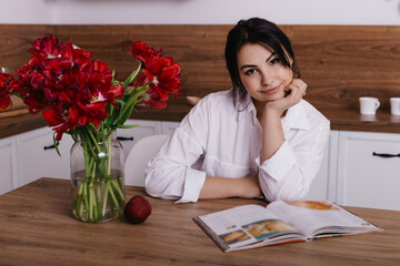 Woman poses with vibrant bouquets of red tulips in vase in kitchen, look and read recipe book. Spring International Women`s Day, 8th of March. Holiday, festive, celebration
