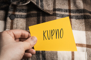 Yellow card with a handwritten inscription "Kupno", a shaft in the hand, protruding from a brown plaid shirt (selective focus), translation: Purchase