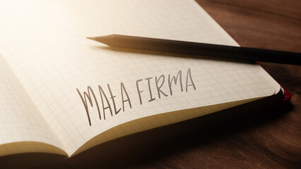 A handwritten inscription "Mała firma" on a grille of an open notebook on a wooden countertop, next to a black pencil, lighting of light. (selective focus), translation: Small company