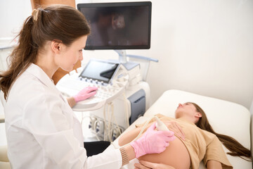 Gynecologist looking at woman while doing ultrasound scanning of pregnant belly