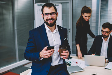Portrait of beautiful attractive young business man in eyeglasses with beard is in office with colleagues on background, prosperous man sitting on table drink coffee smiling and looking at camera