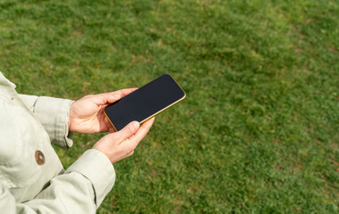 Touch screen phone in hands of woman above the green lawn.