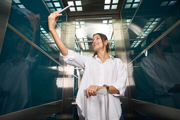 Smiling caucasian woman with suitcase taking selfie on smartphone in elevator