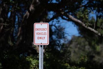 Isolated sign, emergency vehicles only, red and white rectangle with blue skies and trees behind. 