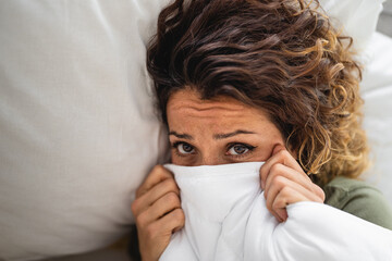 Sad exhausted woman looking at camera wrapped up in a blanket up to her nose due to illness and...
