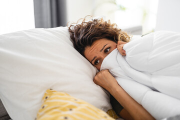 Sad young woman covered in duvet up to her nose lying in cozy bed. She is suffering from heat...
