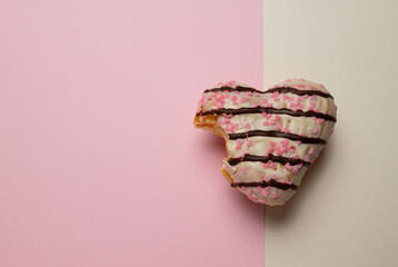 A heart-shaped donut with pink icing and chocolate sprinkles. The donut has been partially eaten,...