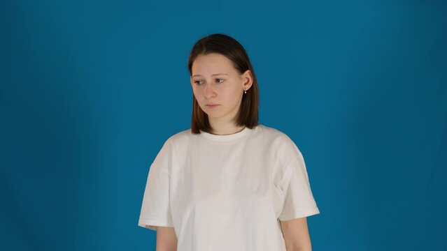 Frustrated young woman in white t-shirt on blue background. Brunette lady suffers from obsessive thoughts in studio. Mental health