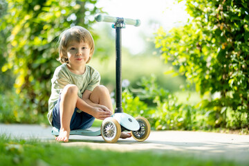 Adorable little boy riding his scooter in a back yard on summer evening. Young child riding a roller. Active leisure and outdoor sports for kids.