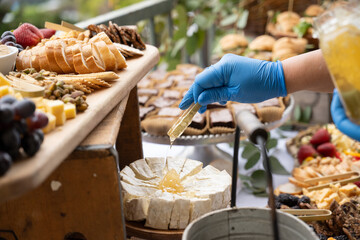 Gloved hand of caterer drizzles honey over dessert cake as finishing touches are made before guest...