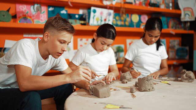 Highschool teenager using carving tool working at clay at pottery workshop. Group of happy diverse children working or modeling cup of clay with dough and equipment scatter around. Edification.