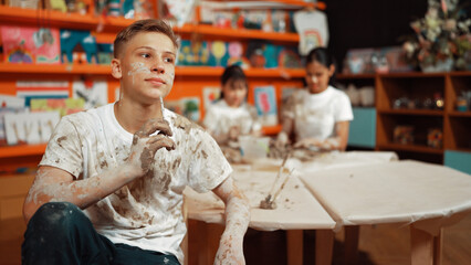 Happy student looking while put paintbrush behind ear in art lesson. Diverse student having pottery...
