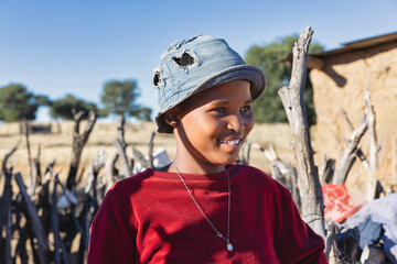 african village, single teenager girl with hat standing in the yard , mud hut and wooden fence in...