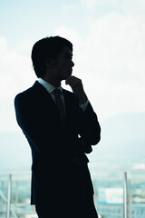 オフィスで腕組みしているスーツ姿の若い男性アジア人ビジネスマン（シルエット・悩む・考える・真剣）
