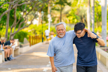 Elderly man suffering knee pain during jogging exercise with his son at park. A man support father...