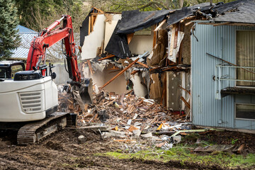 Fototapeta premium Demolishing an old home in a residential neighborhood to clear a lot and make way for a new home, heavy equipment with jawbone bucket to rip apart structure. 
