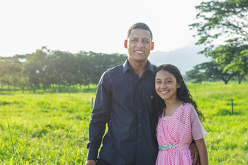 Latino father with his little brunette daughter, standing in a large green field with a sunset in the background.