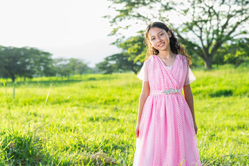 little brunette latina girl in pink dress standing on a grassy field, looking and smiling