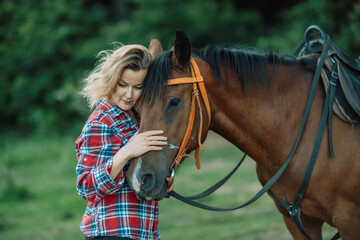 Obraz premium Happy blonde with horse in forest. Woman and a horse walking through the field during the day. Dressed in a plaid shirt and black leggings.