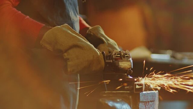 African American blacksmith in apron and protective gloves polishing steel workpiece with angle grinder producing sparks during metalwork in the workshop