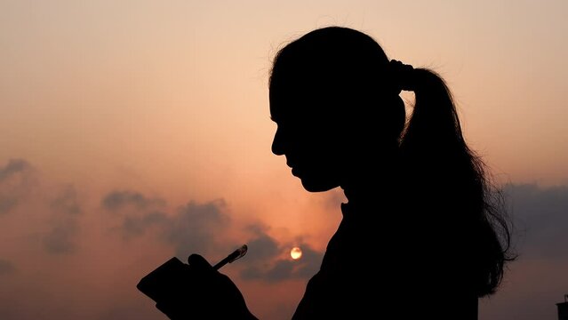 A woman stands under the hot afternoon sun and writes something using a pen on a notepad. A female poet writing concept.
