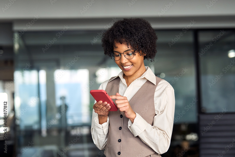 Wall mural Happy smiling busy young professional African American business woman holding cellphone using mobile apps looking at mobile cell phone tech with smartphone in hands standing in office at work.