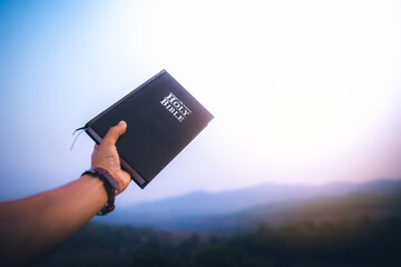 raised hand holding the holy Bible with blue sky in morning. copy space.
