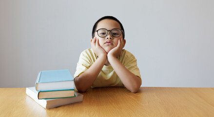 The glasses-wearing child sits with their chin resting on their hands beside a stack of books....