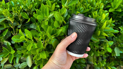 Close up of hand holding ribbed black disposable coffee cup with lid against vibrant green boxwood...