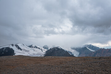Dramatic misty top view from stone pass to big glacier tongue among sharp rocks and large snow-capped mountains in rainy low clouds. Dark atmospheric mountain silhouettes in rain in gray cloudy sky.
