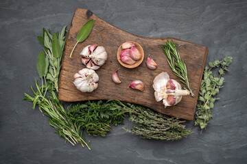 Still life, garlic, fresh green herbs on a rustic wooden cutting board on a black slate plate