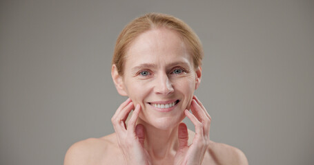 Real people age, beauty, health and dry skin care concept - beautiful mature Caucasian middle aged woman in her 50s touching facial skin doing facial massage and looking at camera