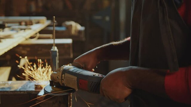 African American blacksmith in apron and protective glasses polishing steel billet with angle grinder producing sparks at workbench in workshop. Close-up view
