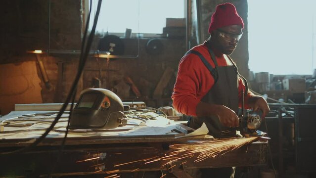 African American blacksmith in apron and safety glasses cutting ferrous metal with angle grinder producing sparks during the day at his workshop