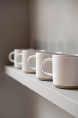 A series of minimalist ceramic or porcelain mugs with clean lines and matte finishes, arranged in a row on a solid-colored shelf, inviting relaxation and indulgence with every sip. 