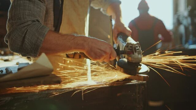 Hands of professional blacksmith in apron shaping metal detail with angle grinder tool producing sparks during metalwork. Close-up view