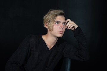 A young attractive guy is sad while sitting on a chair. Black background.