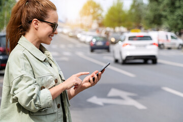 Urban woman using her smartphone while standing next to city road. Taxi online service. Mobility as...