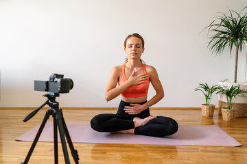 Fitness coach records video of meditation breathing exercise while sitting on yoga mat at home.