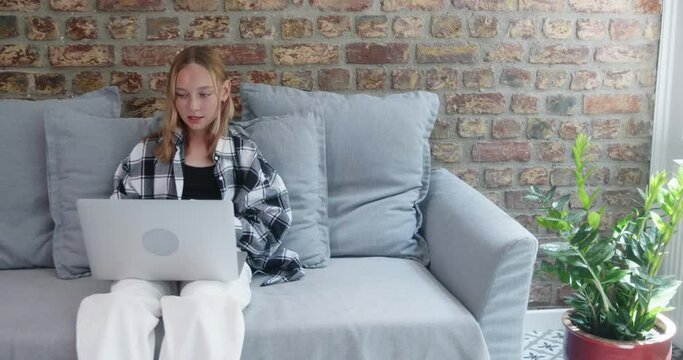 Teenager gesturing while reading online on laptop. Girl in checkered pattern casuals sitting on sofa against wall. She is spending leisure time at home during sunny day. 