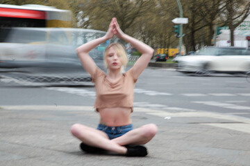 Junge Frau mit langen blonden Haaren sitzt auf dem Gehweg und meditiert. Um sie herum rasender Verkehr unscharf fotografiert
