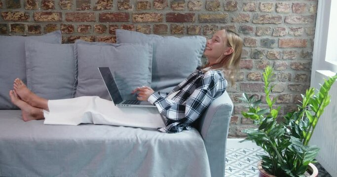 Cheerful teenage girl laughing while using laptop. Thoughtful female with wireless computer sitting on cozy sofa. She is relaxing in living room at home.