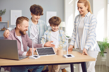 Happy smiling young family learning about sustainable renewable energy sitting at table in living room at home with children looking into wind turbine and solar panel models. Green energy concept.