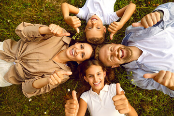 Top view of happy young family of four lying on green grass and showing thumb up sign in summer park. Smiling parents having fun with kids boy and girl in nature enjoying time together.
