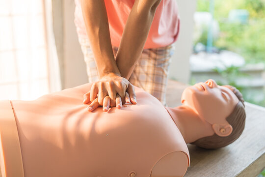 Close-up of young Asian female hands performing cardiopulmonary resuscitation (CPR) on a training dummy. Key elements include defibrillator, compressions, mouth-to-mouth, and abdominal thrusts.