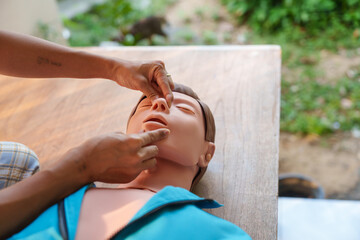 Close-up of young Asian female hands performing cardiopulmonary resuscitation (CPR) on a training dummy. Key elements include defibrillator, compressions, mouth-to-mouth, and abdominal thrusts.